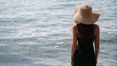 A picture of a beautiful young woman with long dark hair in a dress and a straw hat, posing on the beach, standing with her back and looking at the endless sea. A place for your text.の写真素材