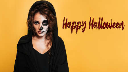 Bloody HAPPY HALLOWEEN text on a yellow background. Next to a girl in a hood with scary makeup. The concept of a costumed party.の写真素材