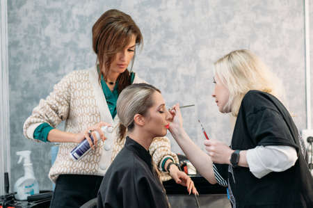 A hairdresser and a makeup artist together do styling and makeup for a young and beautiful model girl in a beauty salon.の写真素材