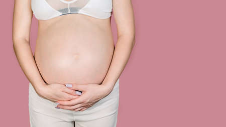 A young adult pregnant woman in white clothes touches her big naked belly with her hands. Showing the form. The concept of pregnancy. Close-up. Isolated on a pink background. A place for your text.の写真素材
