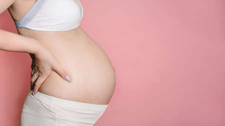 A young adult pregnant woman in white clothes touches her big naked belly with her hands. Showing the form. The concept of pregnancy. Close-up. Isolated on a pink background. A place for your text.の写真素材