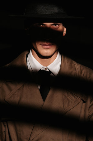 A dark portrait of a male detective in a coat and hat. Spectacular light from behind the blinds.. Dramatic noir silhouette in the style of detective spy films and books of the 1950s.の写真素材