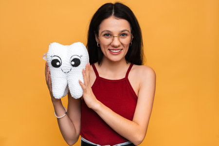 A smiling young woman with healthy teeth holds a white plush tooth on a blue isolated background. Oral hygiene. Prosthetics. Rules for brushing teeth.の写真素材