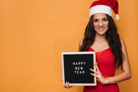 A smiling Caucasian woman in a red Santa hat holding a letter board with the inscription Happy New Year on it. On a yellow isolated background. A place for your text.の写真素材