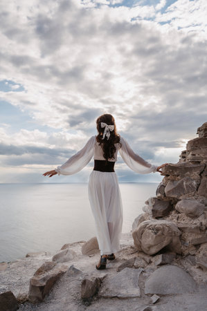 A young woman in a white dress stands barefoot on a cliff face in full height. The dress flutters in the wind. A sacred glow breaks through the clouds. The girl looks like an angelの写真素材