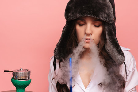 An attractive woman in the traditional Russian hat with earflaps and glasses smokes a hookah and enjoys smoking. Pink solid color background.の写真素材
