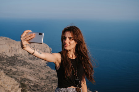 A young girl takes a selfie on her smartphone against the background of the sea at the cliff. The girl smiles at the camera. The concept of tourism, recreationの写真素材