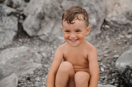 The child boy is sitting, smiling and sunbathing on large boulders, on the beach near the sea.の写真素材