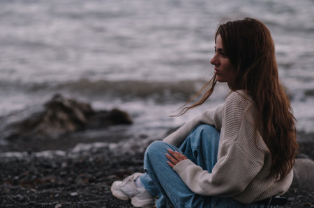 A young Caucasian girl is sitting on a rock by the seashore, with a thoughtful look. Gray, cool, inclement weather.の写真素材