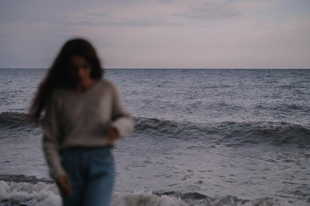 A young Caucasian girl walks along the coastline of the sea, and looks at her feet. A blurred image. Gray, cool, inclement weather.の写真素材