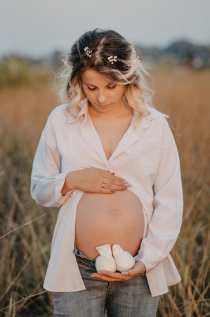 A young expectant mother in the third trimester of pregnancy in a white dress hugs her stomach and holds booties against the background of the natural landscape. The concept of future motherhood.の写真素材