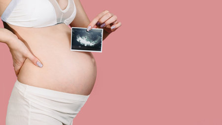 A happy expectant mother shows an ultrasound image of the fetus on camera. Prenatal ultrasound screening. close-up of the abdomen of a young pregnant woman holding a sonogram of an unborn child. A place for your text.の写真素材