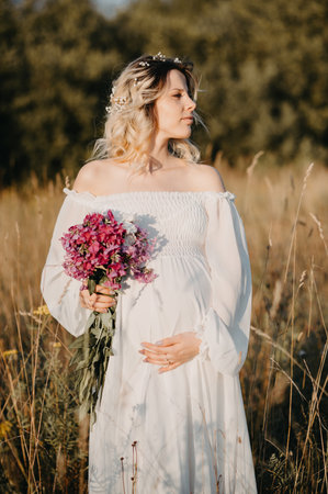 A young expectant mother in the third trimester of pregnancy with a bouquet of flowers in her hands, in a white dress hugs her stomach against the background of a natural landscape. The concept of future motherhood.の写真素材