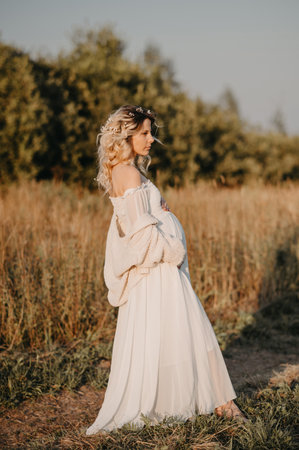 A young expectant mother in the third trimester of pregnancy in a white dress hugs her stomach against the backdrop of a natural landscape. The concept of future motherhood.の写真素材
