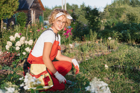 A pretty female gardener takes care of plants in her garden. The gardener moves the soil with a shovel, prepares it for planting. Gardening and floriculture. Growing flowers in the home garden.の写真素材