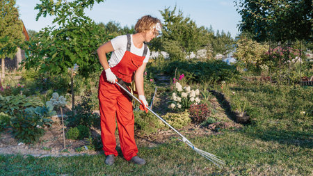 A happy female gardener in gloves and an apron stands against the background of a flower bed in a home garden with a rake in her hands. Gardening and floriculture. Flower careの写真素材