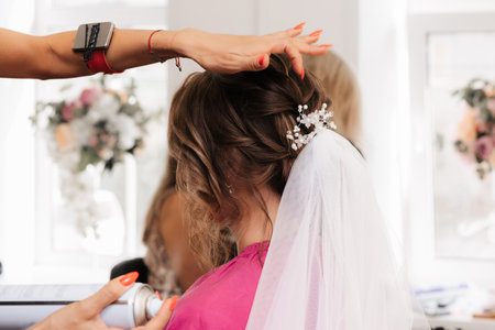 A hairdresser makes an elegant hairstyle for styling a bride with a white veil in her hair in the salonの写真素材