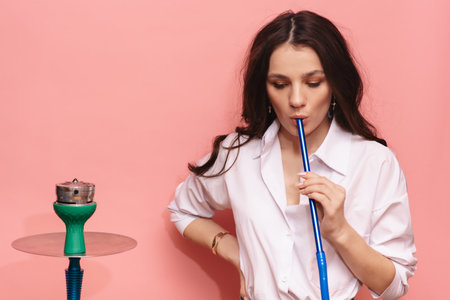 Young woman smokes a hookah, sheesha on a pink background. The pleasure of smoking.の写真素材