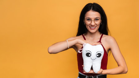 A smiling young woman with healthy teeth holds a white plush tooth on a blue isolated background....