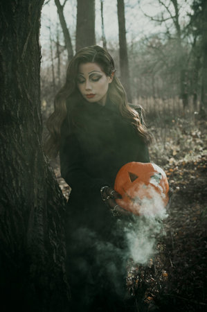 A young woman in a Gothic gloomy image of a witch surrounded by smoke, a pumpkin in her hands. Halloween costume. Vertical photo.の写真素材