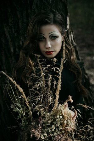 Portrait of a young woman in the Gothic gloomy image of a witch in the forest with a bouquet. Halloween costume. Vertical photo. Blur.の写真素材