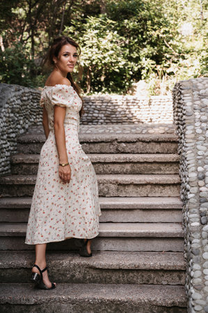 A young girl, a brunette with long hair, in a vintage dress is standing on the steps of a gray stone staircaseの写真素材