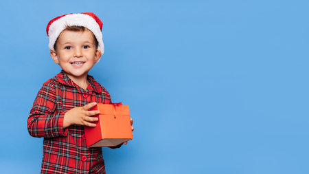 A happy little boy in Christmas pajamas and a hat with a gift box. A place for your text. Studio shot isolated on a blue background. The concept of holidays.の写真素材