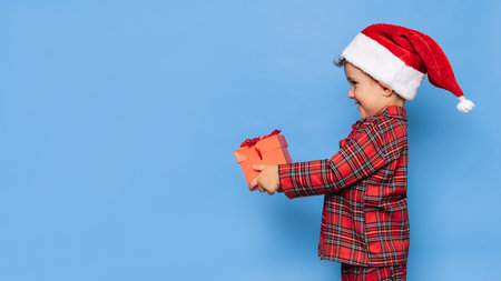 A smiling little boy in Christmas pajamas and a hat with a gift box. A place for your text. Studio shot isolated on a blue background. The concept of New Year and Christmas.の写真素材