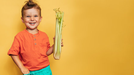 Caucasian little happy boy holds a fresh raw celery stalk on a yellow background. Space for text. Healthy lifestyle, diet concept, vegetarianism, raw food.の写真素材