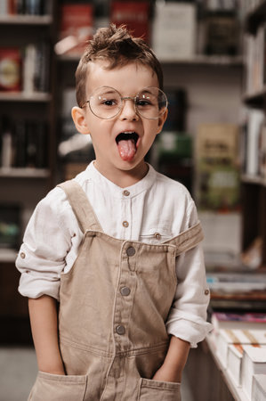 A cute Caucasian kid, a boy with round glasses, is fooling around in a bookstore among the shelves with books.の写真素材