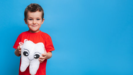 A smiling boy with healthy teeth holds a plush tooth in his hands on a blue isolated background. Oral hygiene. Pediatric dentistry. Prevention of caries. A place for your text.の写真素材