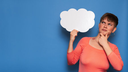 Studio shot of a young woman with a puzzled expression, holding a white empty banner in the form of a cloud on a blue background. Copy space, space for your ad or text.の写真素材