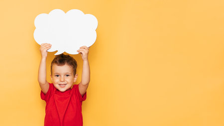 Studio portrait of a happy boy with a clean white board in the shape of a cloud on a bright yellow background, with a place for your text or advertisingの写真素材