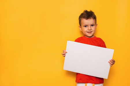 Studio portrait of a happy boy with a clean white poster in his hands on a bright yellow background, with a place for your text or advertisingの写真素材