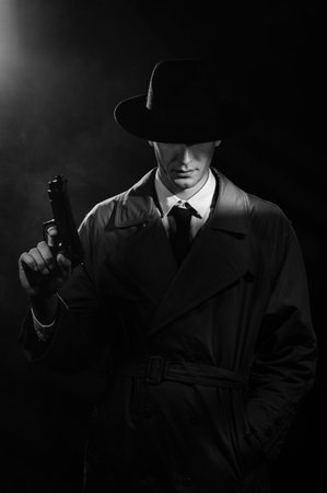 A dark silhouette of a male detective in a coat and hat with a gun in his hands in the Noir style. The head is lowered. A dramatic noir portrait in the style of detectives of the 1950s.の写真素材