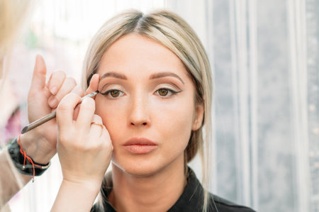Shooting in a beauty salon. A young beautiful girl model master makeup artist puts shadows on the eyelid draws arrows on the eyelid.の写真素材