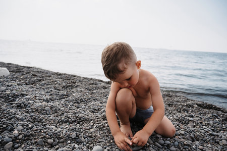 A cute boy is relaxing on the beach, playing with sea pebbles. Outdoor activities on the seashoreの写真素材