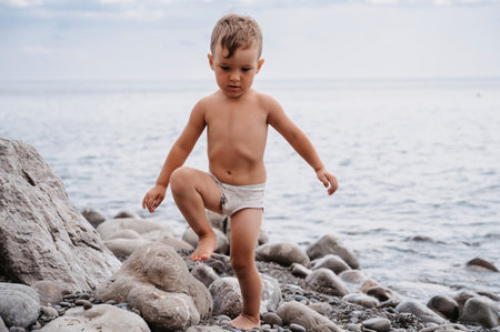 Cute baby boy climbs on rocks, on a beach of pebbles. Active recreation of the coast in the open air.の写真素材