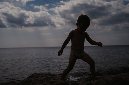 Cute baby boy climbs on rocks, on a beach of pebbles. Active recreation of the coast in the open air.の写真素材