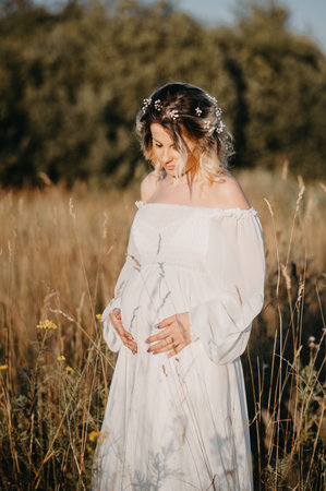 A young expectant mother in the third trimester of pregnancy in a white dress against the background of a natural landscape. The concept of future motherhood.の写真素材