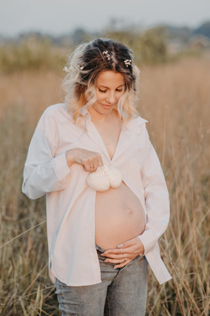 A young expectant mother in the third trimester of pregnancy in a white dress hugs her stomach and holds booties against the background of the natural landscape. The concept of future motherhood.の写真素材