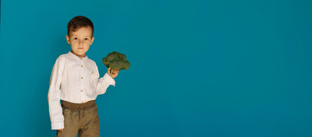 A studio shot of a smiling boy holding fresh broccoli on a blue background with a copy of the space....