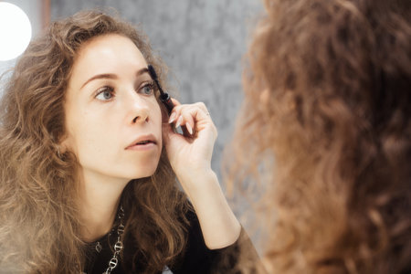 Beautiful girl is doing a makeup, retouching eyebrows while looking at the mirror