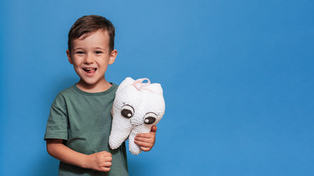 A smiling boy with healthy teeth holds a toothbrush and a plush tooth on a blue background....