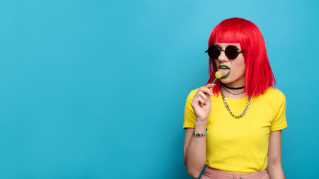 A bright and funny young woman with red hair licks a lollipop. Studio colorful picture of a pin-up style woman with a lollipop on a stick, on a blue background. A place for your text.の写真素材