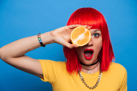 A young beautiful funny model with a slice of orange near her eyes, in the background with makeup, hairstyle. Bright, funny summer photo. Bright background.の写真素材