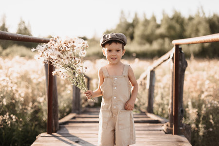 A boy is playing on a farm or ranch field, resting. Portrait of a child boy in a cap and overalls standing on the bridge. Childhood.の写真素材