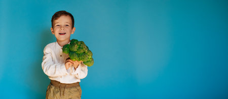 A studio shot of a smiling boy holding fresh broccoli on a blue background with a copy of the space. The concept of healthy baby foodの写真素材