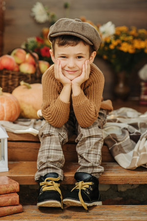 A Caucasian boy in a sweater sits on the porch of his house decorated with pumpkins in the autumn...