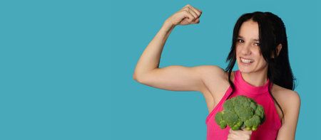 A smiling white girl with fresh broccoli in one hand and showing off her biceps in the other. Panoramic banner, free space.の写真素材
